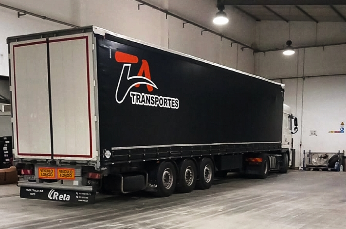 Imagem do serviço de carga completa, um camião virado com caixa preta com logotipo da Ta Transportes, dentro do armazém.